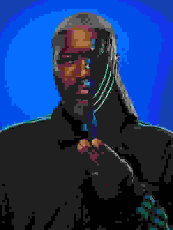 A man with a beard and braided hair wearing a black jacket against a vibrant blue background.