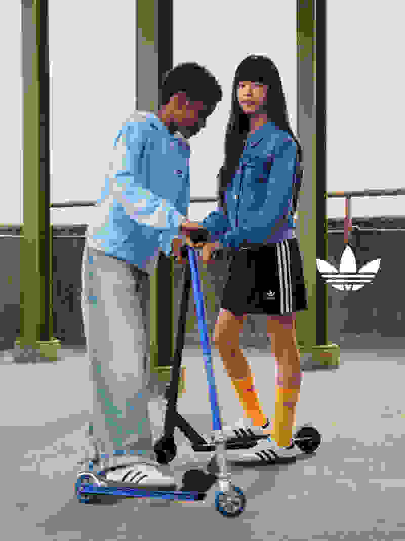 A girl and boy wear white adidas Samba shoes while posing with a scooter on a concrete street.  