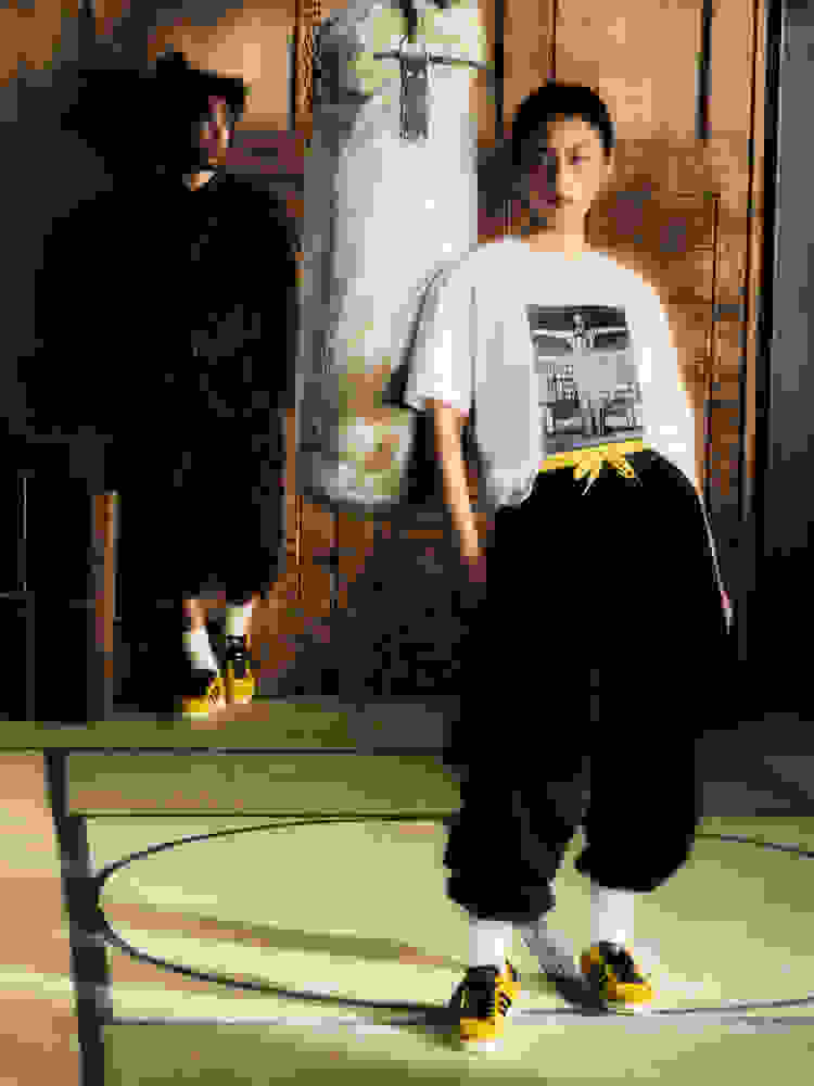 Two people wearing yellow adidas Originals x Bruce Lee Jabbar Lo sneakers with black stripes, standing in a wooden room with a hanging punching bag.