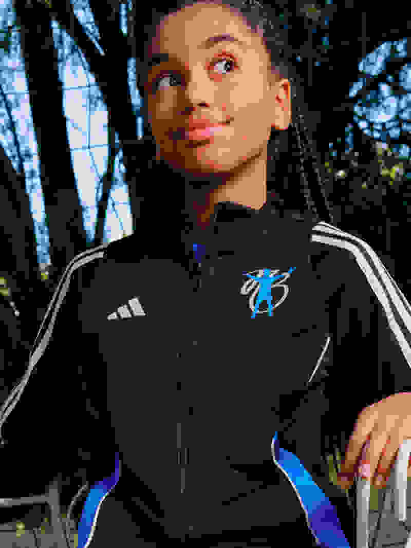 A young girl with long black braids smiles brightly in a black tracksuit from the adidas Bellingham kids collection.