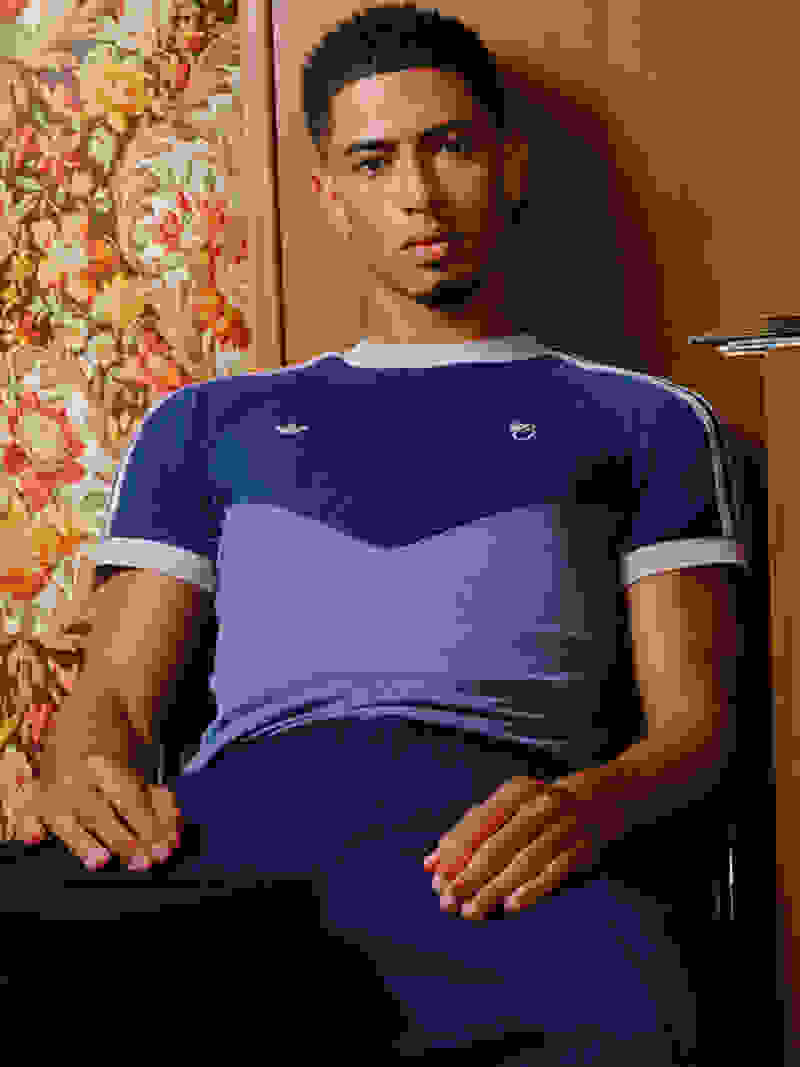 Jude Bellingham leans casually against a wall, fully dressed in the adidas Bellingham collection, looking straight into the camera.