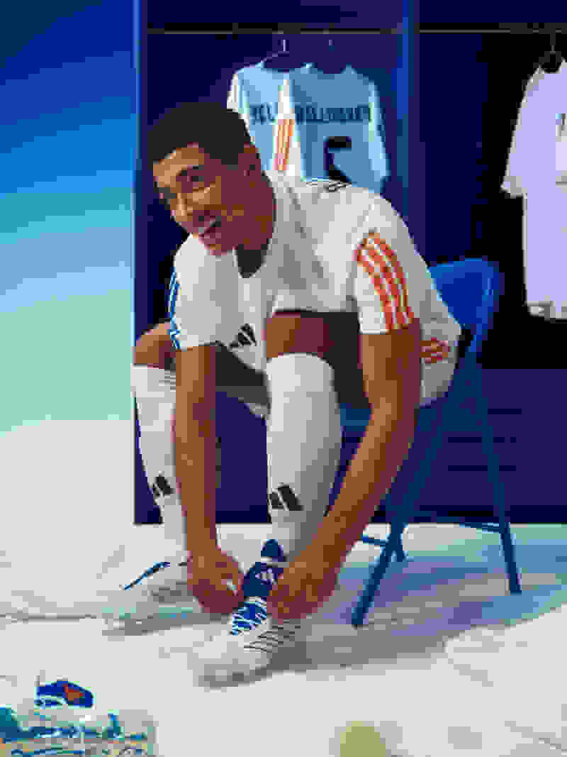 Jude Bellingham ties his red, white, and blue adidas Predator boots while seated in full match kit on a blue chair.