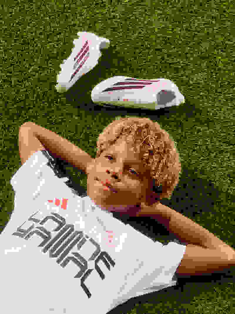 Um rapaz loiro está no relvado ao lado das botas F50 de Lamine Yamal, braços atrás da cabeça, a sorrir para o céu.