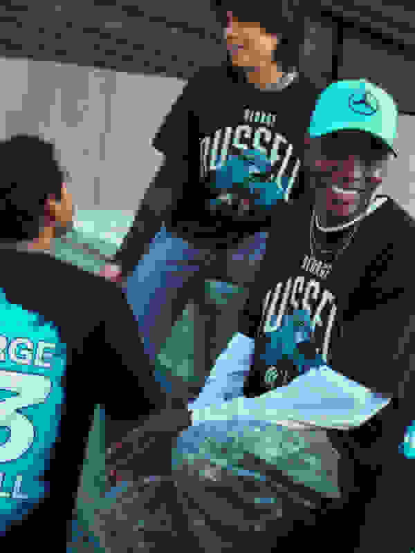 Two young individuals, one wearing a "George Russell" t-shirt and the other a "Mercedes" cap, are engaged in a friendly conversation in what appears to be an outdoor setting with a metal structure in the background.