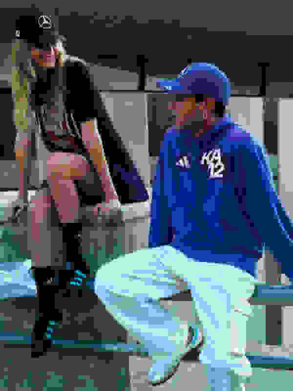 Two individuals, a woman wearing a black shirt and a man wearing a blue sweatshirt and cap, are seated on a bench in what appears to be an indoor setting with glass walls or windows in the background.