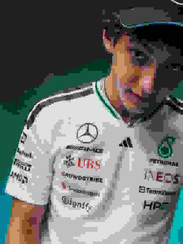 A young man wearing a white racing uniform with various sponsor logos, standing against a dark background.