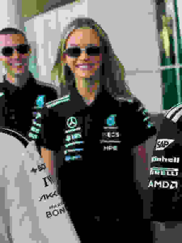 Fotoğraf, üzerinde çeşitli sponsor logoları bulunan siyah bir yarış forması giyen ve bir yarış arabasının önünde duran gülümseyen bir kadını gösteriyor.