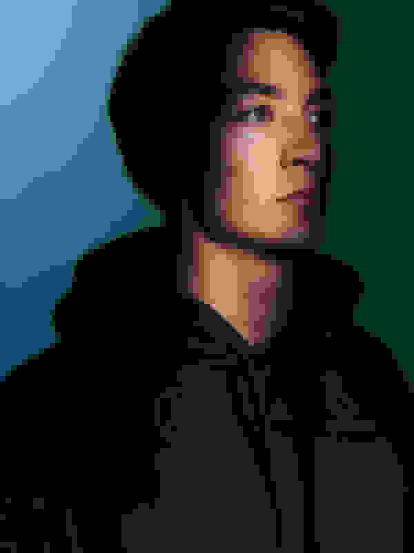 A young man with a serious expression, wearing a black hooded sweatshirt, set against a teal-coloured background.