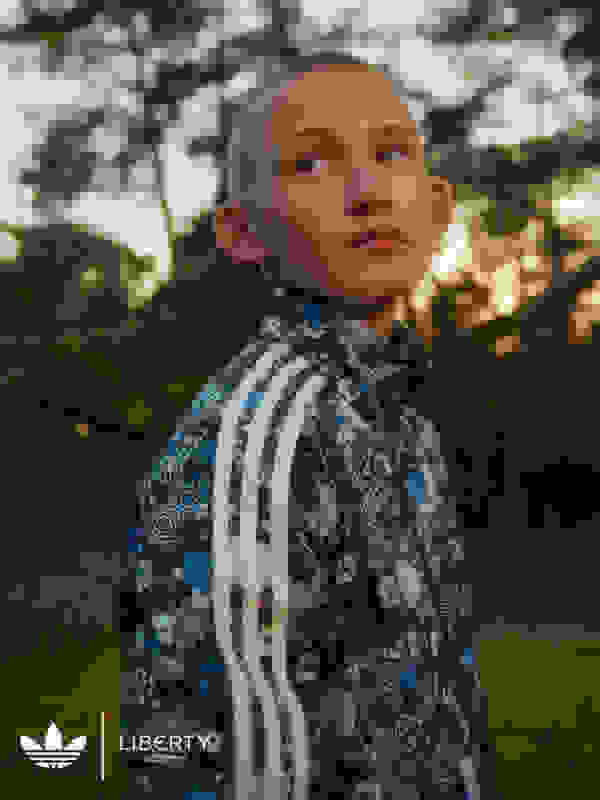 A boy outdoors with trees behind him, wearing a navy blue adidas x Liberty London jacket printed with flowers and white 3-Stripes.