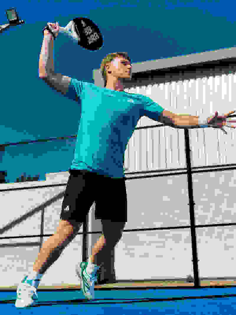 A person in a green shirt and black shorts is playing padel tennis on an outdoor court with a blue sky in the background.