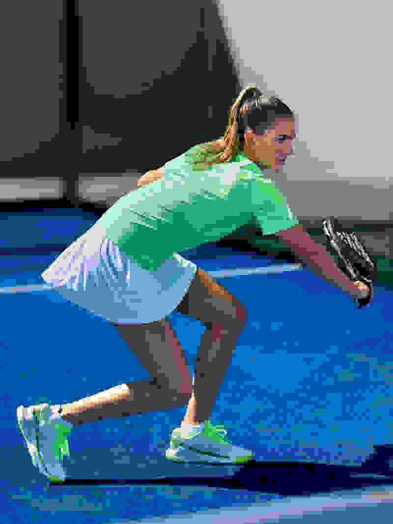 A female athlete in a bright green top and white shorts is crouched on a blue padel court, preparing to make a move.