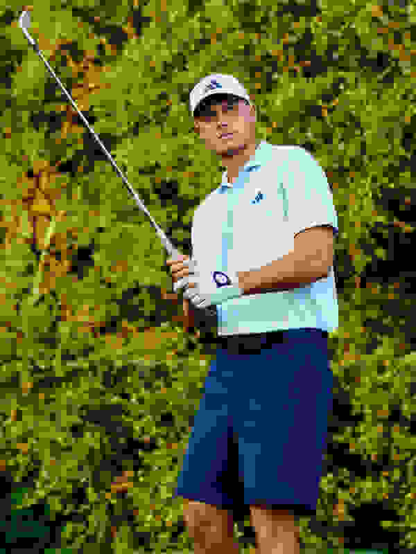 Een man in een lichtblauw poloshirt en een marineblauwe short houdt een golfclub vast en staat in een weelderige, groene outdooromgeving.