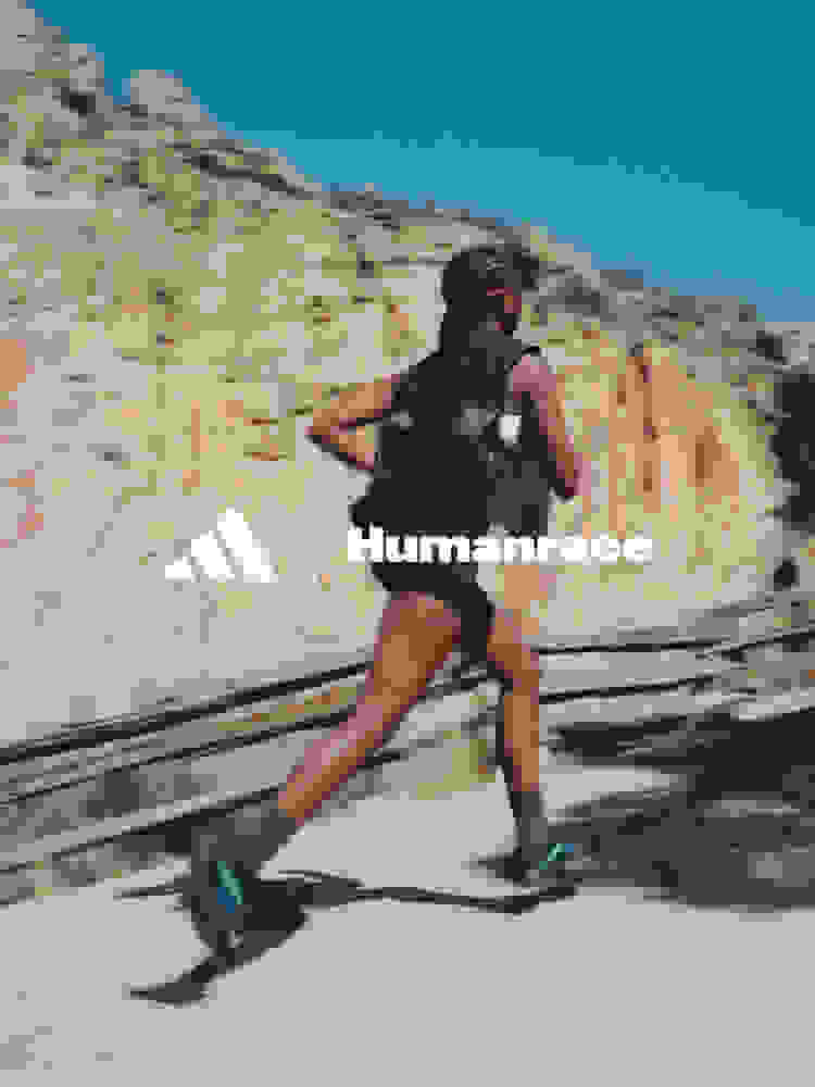 A person wearing a black vest and running on a rocky, desert-like terrain with mountains in the background, against a clear blue sky.