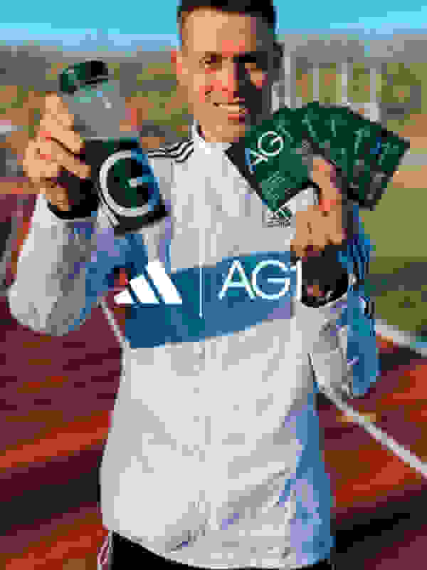 A smiling young man in an Adidas jacket holds a bottle of AG1 supplement and various AG1 packets in front of a red running track.