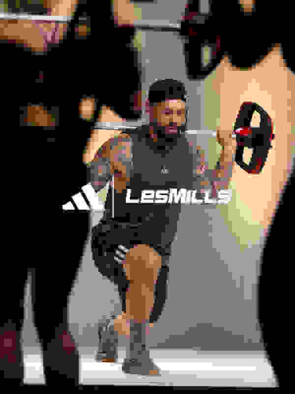 A muscular man with tattoos and a beard is performing a workout exercise while wearing Lesmills branded athletic gear, with a dark background and other people visible in the background.
