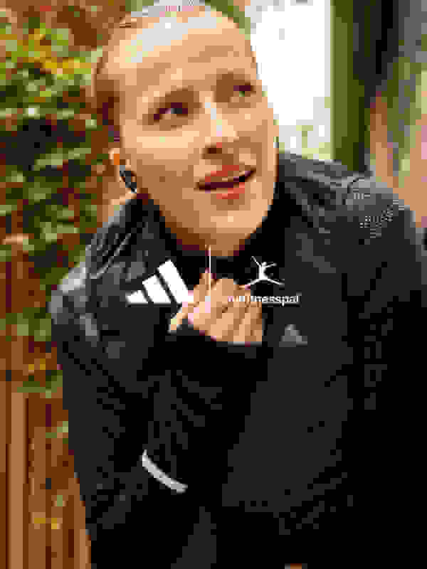 Eine lächelnde Frau in einer schwarzen Sportjacke mit dem „myfitnesspal“-Logo steht in einer grünen Outdoor-Kulisse.