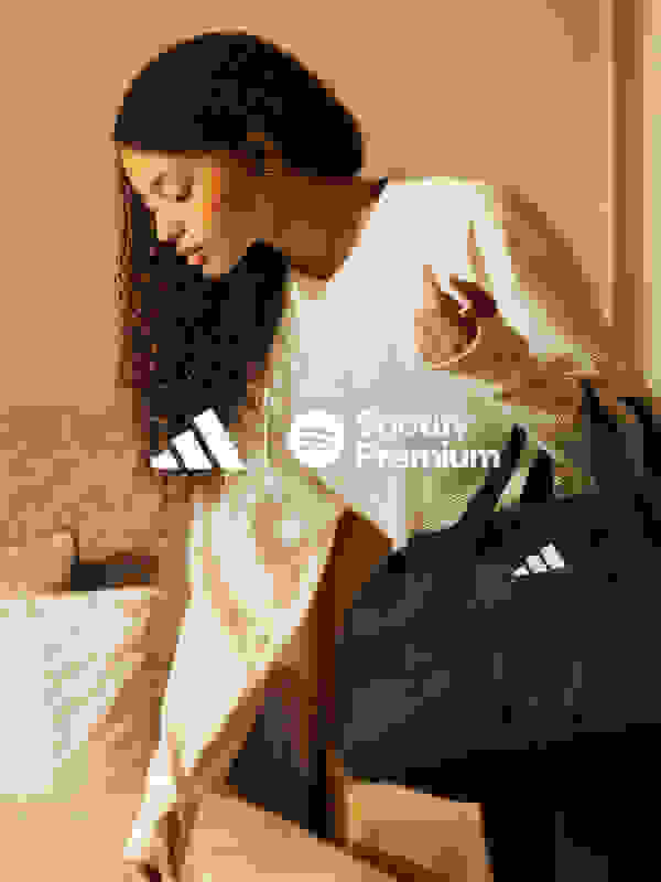 Eine junge Frau mit lockigem, dunklem Haar in einem weißen Spotify Premium Sweatshirt sitzt in einer schwarzen Sporttasche auf einem Bett.