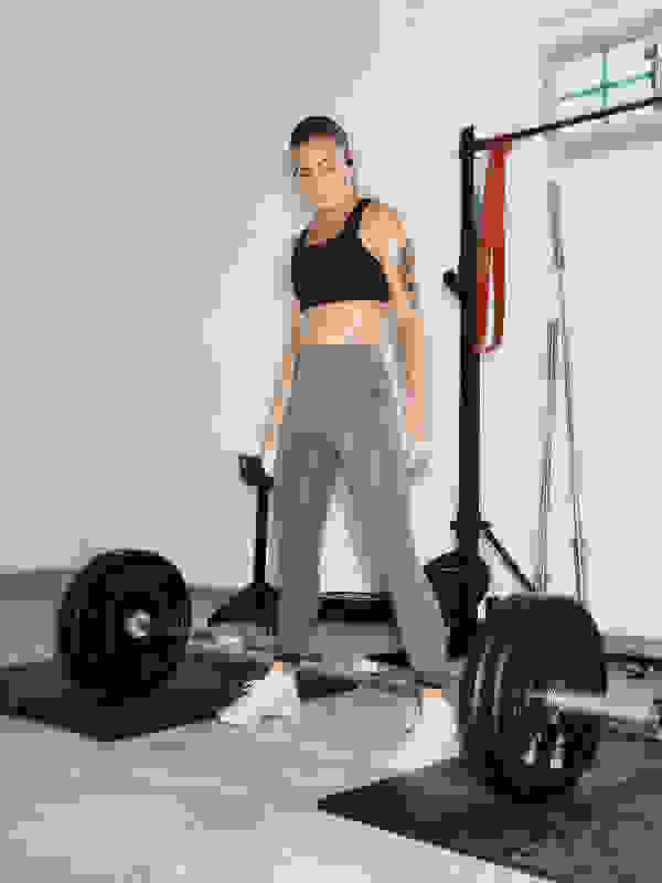 Uma mulher em roupa atlética destaca-se num ambiente de ginásio, com equipamento de exercício e uma barra visível ao fundo.