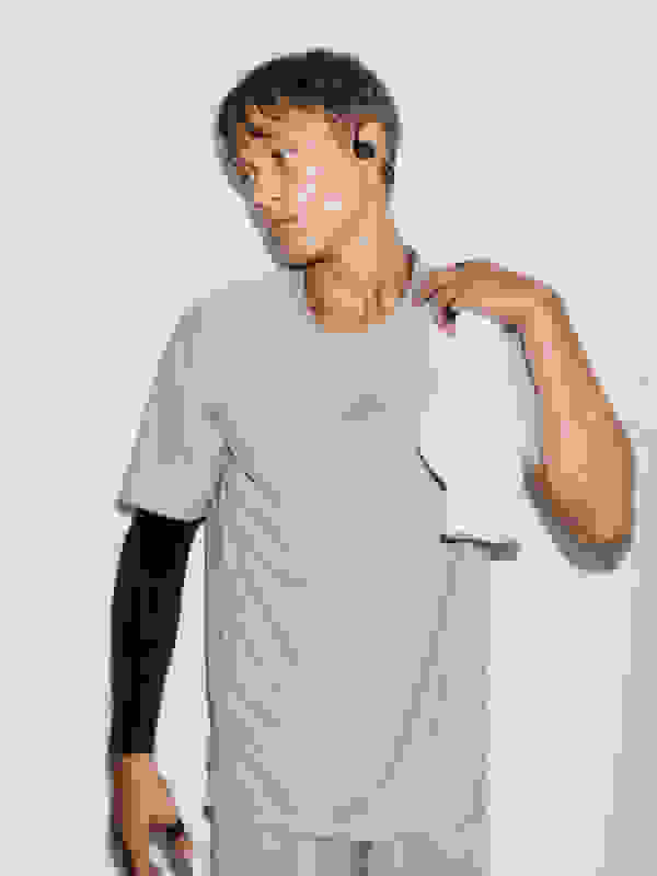 Một thanh niên mặc áo sơ mi thể thao màu xám và quần short đen đứng trên nền trắng trơn, cầm khăn trắng.