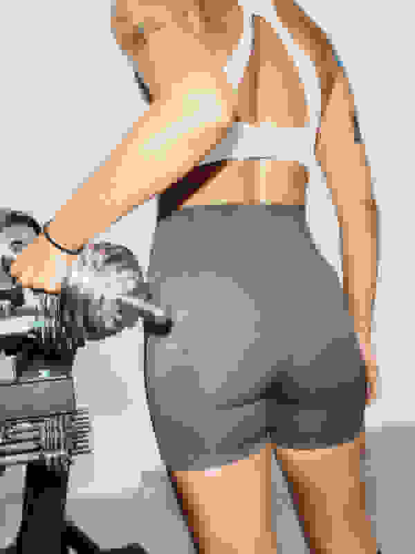 Hình ảnh này cho thấy phần thân dưới và ống chân của một người, mặc quần short màu xám bó sát, đứng cạnh một mẩu trang thiết bị tập luyện.