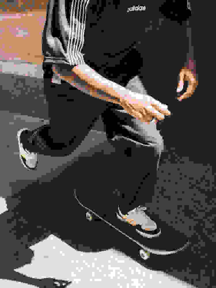 A person wearing an Adidas shirt and black pants is crouching on a skateboard on a concrete surface.