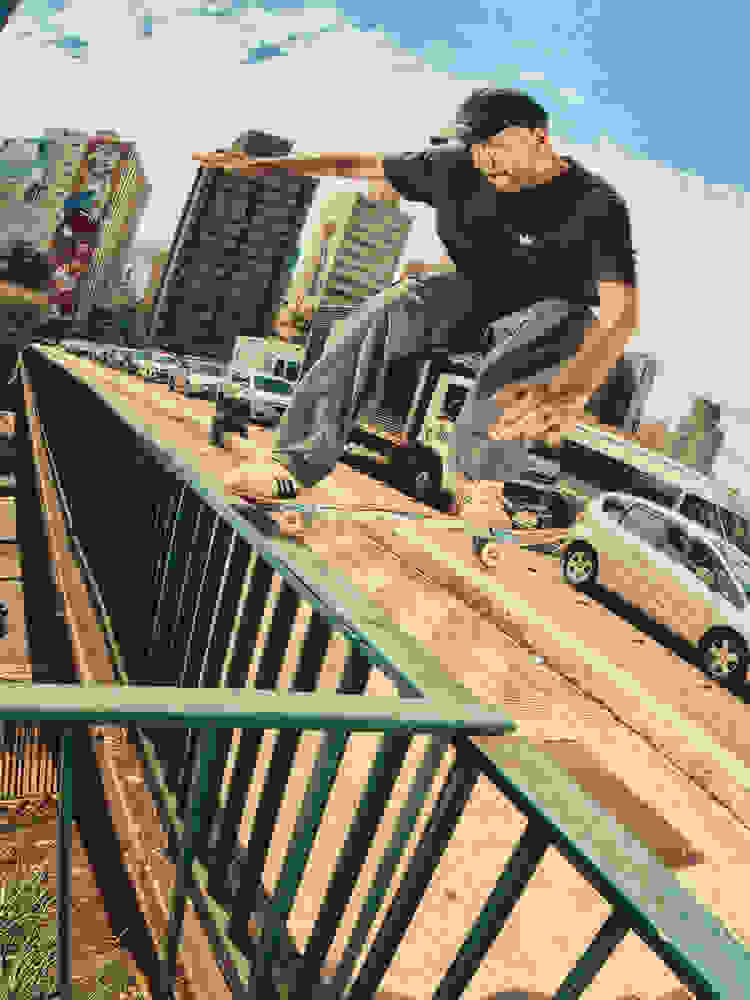 A man skateboards along a handrail above a busy city street, wearing a black adidas t-shirt with the Trefoil logo, light grey pants, and beige adidas Glenburn sneakers with black stripes.