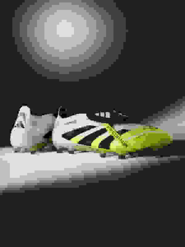 A pair of white and neon yellow soccer cleats are prominently displayed against a dark, shadowy background, creating a dramatic and striking visual composition.