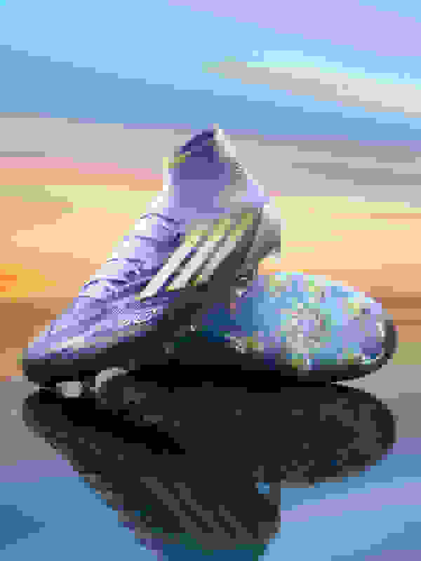 A pair of vibrant purple and green soccer cleats are reflected in a calm, reflective surface against a colorful sunset sky.