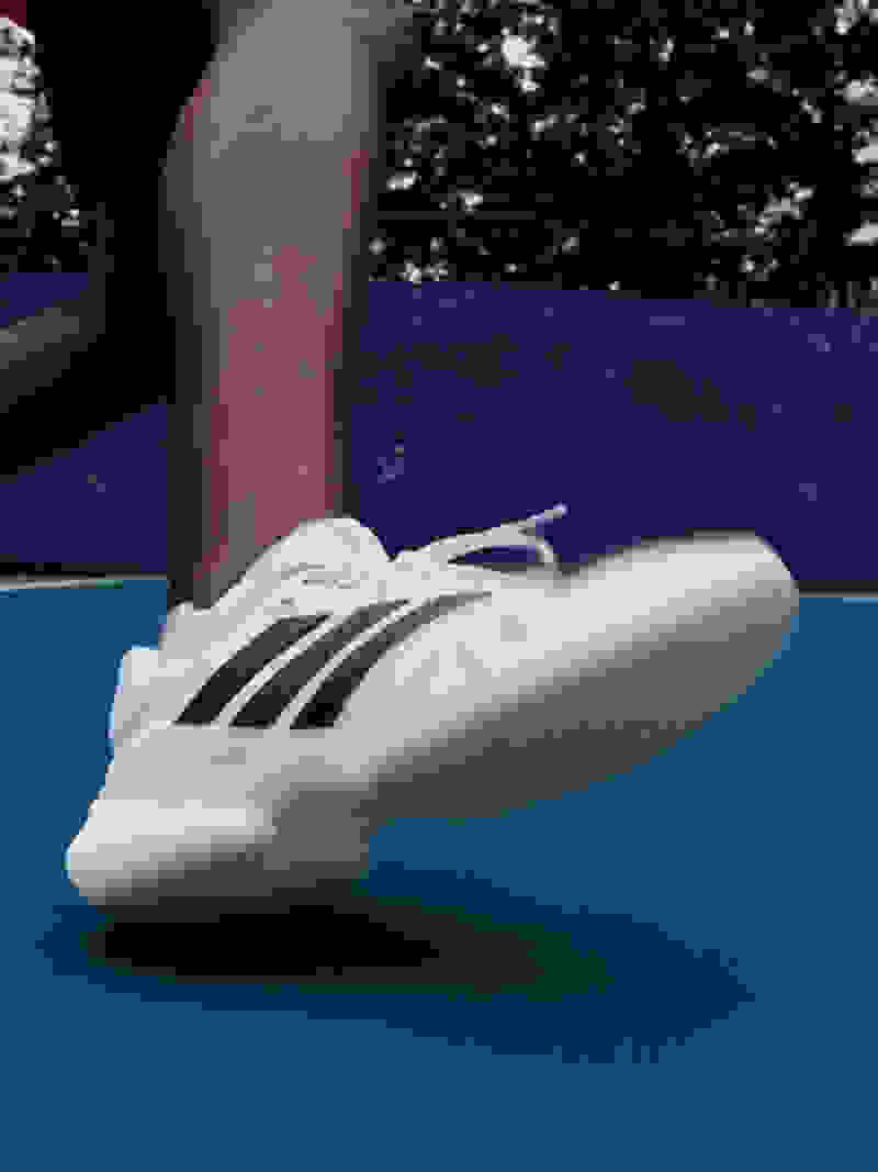 Hình ảnh này cho thấy bàn chân của một người đang mang đôi giày thể thao màu trắng với các sọc đen trên bề mặt màu xanh dương, có thể là sân tennis, với các cây cối nhìn thấy phía sau.