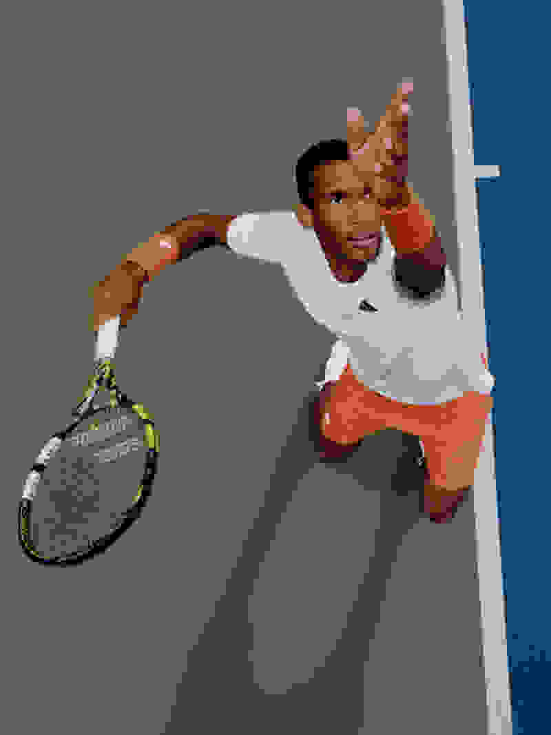 Vận động viên quần vợt mặc áo thun trắng và quần short màu cam là người bạn đồng hành giữa các cú vung vợt, với vợt tennis cầm tay trên sân tennis màu xanh.