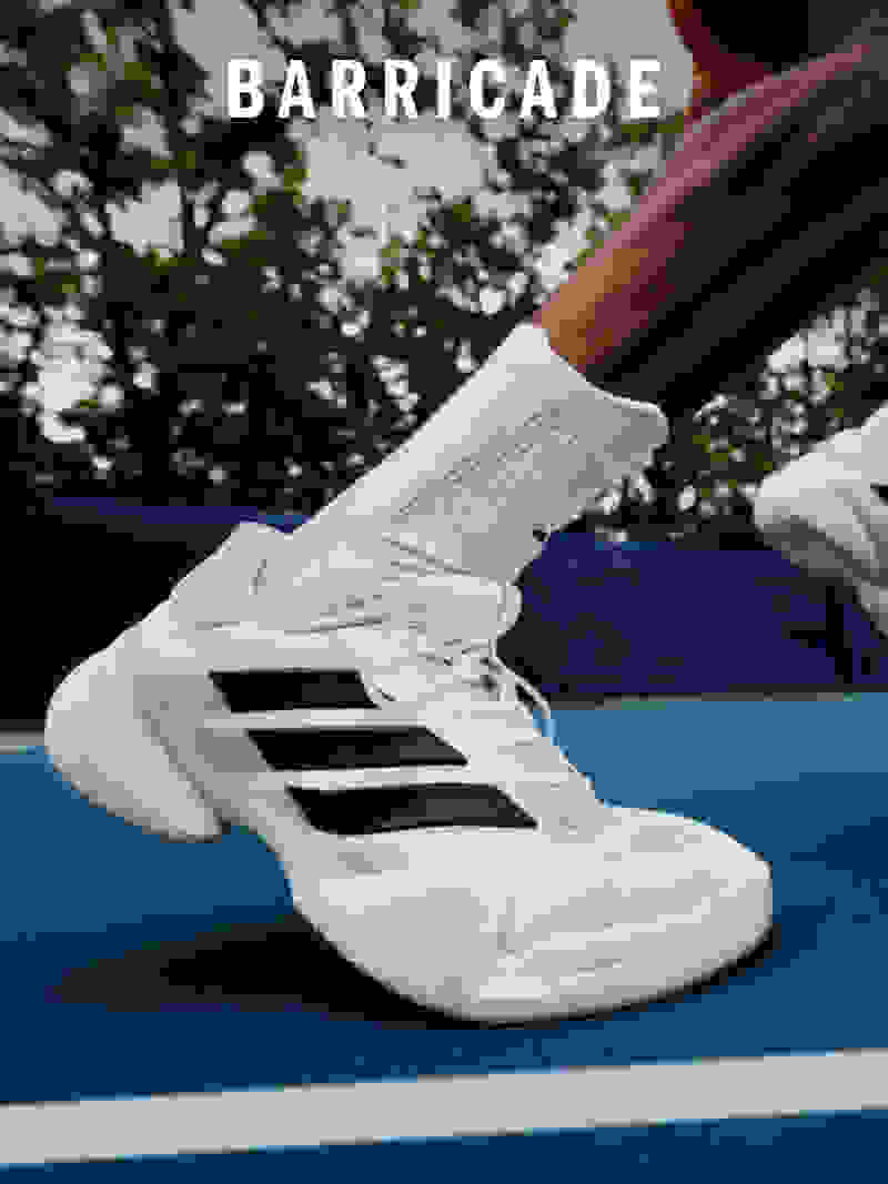 L'immagine mostra un paio di scarpe sportive bianche e nere con la parola "BARRICADE" in evidenza sul lato. La scarpa è posizionata su una superficie blu, con uno sfondo naturale sfocato di alberi e foglie visibile.