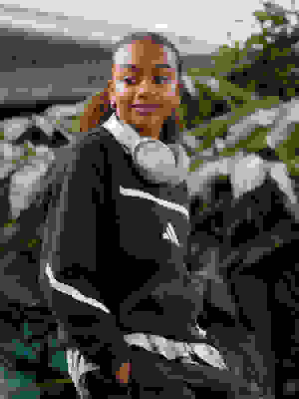 A young woman with curly hair wearing a black and white athletic jacket stands in a lush, green outdoor setting.