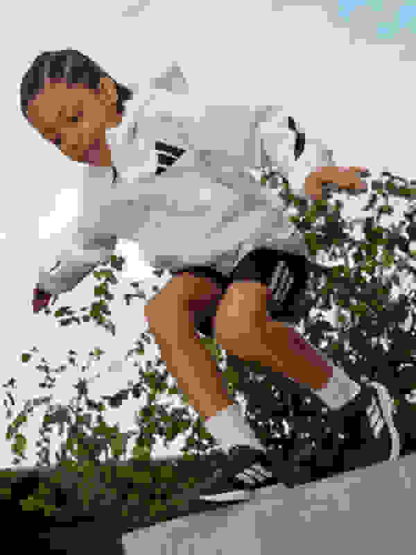 A person in a white athletic jacket and black shorts is jumping over a low wall, surrounded by greenery in the background.
