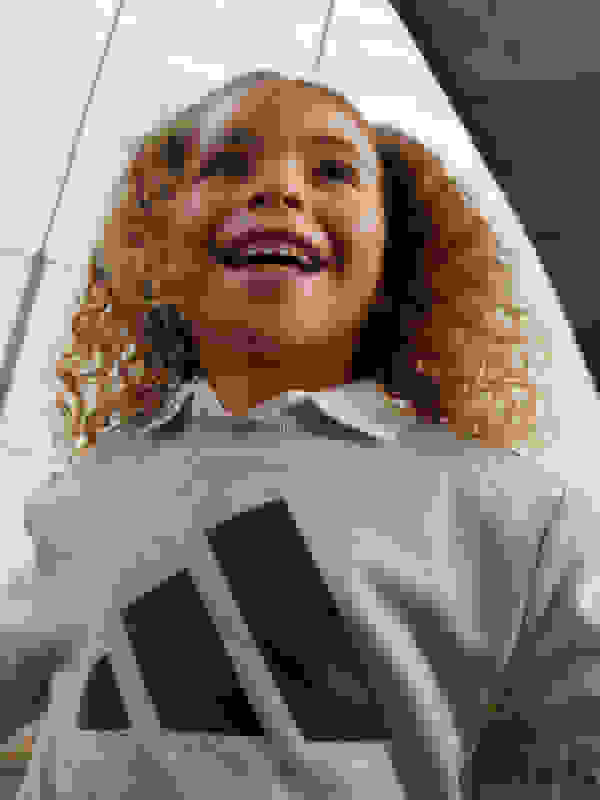 A young girl with curly hair and a bright smile, wearing a gray sweatshirt with the Adidas logo, stands against a white background.