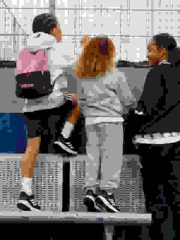 Three young individuals, likely students, standing together and interacting in what appears to be a school or educational setting, with a wire mesh fence visible in the background.