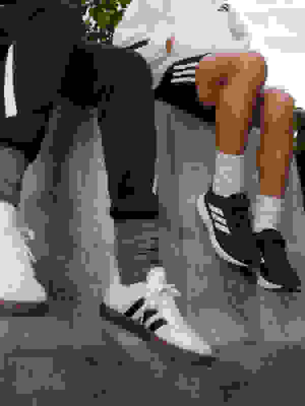 The image shows the lower body of two people sitting on a grassy surface, with one person wearing black pants and the other wearing white pants. The background appears to be a natural setting with some foliage visible.