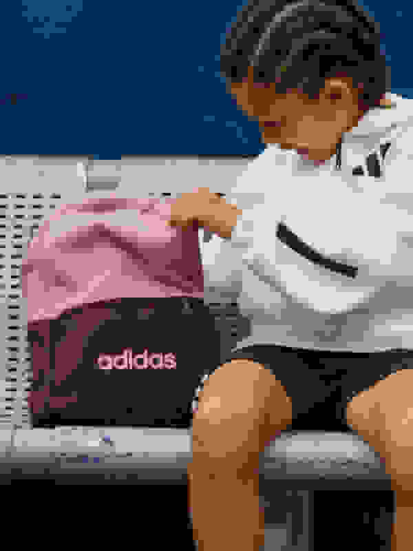 A young child, wearing a white athletic jacket, sits on a bench and examines the contents of a pink and burgundy Adidas bag.