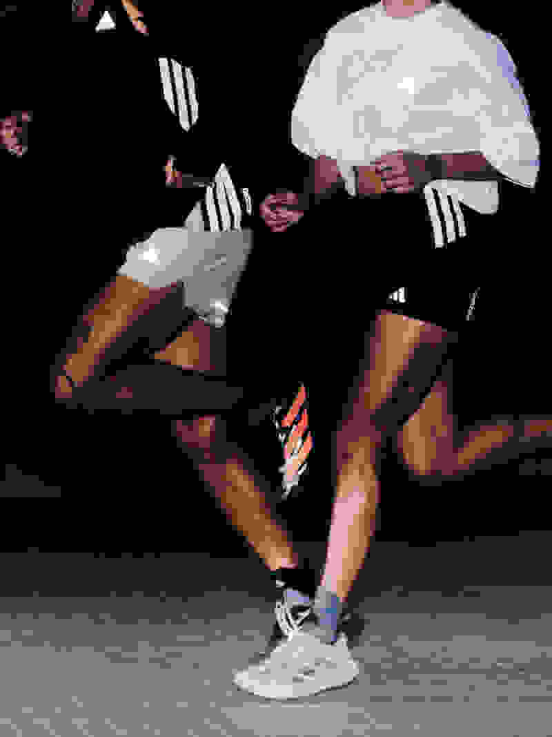 The image shows two individuals wearing white and black athletic clothing, engaged in what appears to be a physical activity or performance on a dark background.