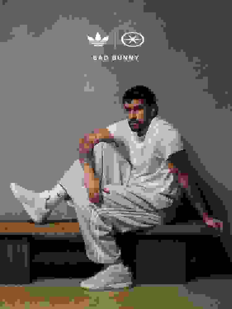 Bad Bunny olha para a câmera, sentado em um banco de madeira sobre uma parede cinza texturizada, vestindo uma camiseta branca, calça esportiva adidas cinza claro, meias adidas brancas e tênis adidas branco. Uma perna se flexiona no banco e o outro pé repousa no chão.