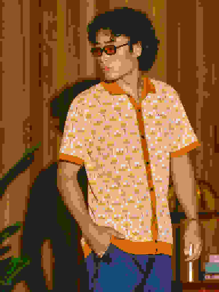 Člověk s kudrnatými vlasy a brýlemi v vzorované žluté košili stojí v místnosti s dřevěnými stěnami a květinovou květinou v pozadí.