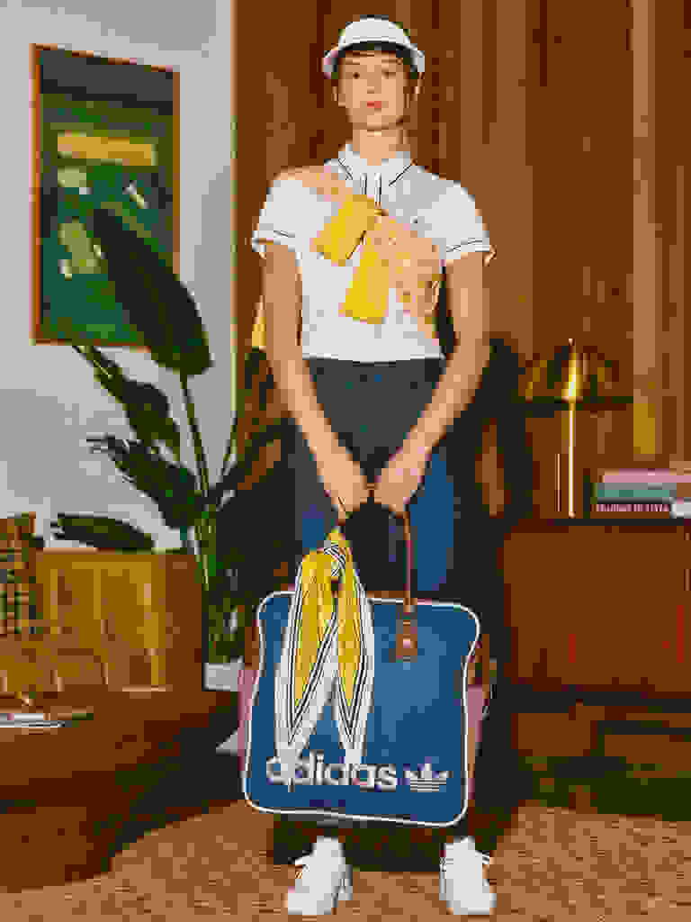 Mladá žena v bílém tričku, námořnicky modré sukni a bílé čepici stojí v místnosti se zelenou kresbou na zdi, květinovou květinou a dřevěným stolem s lampou.