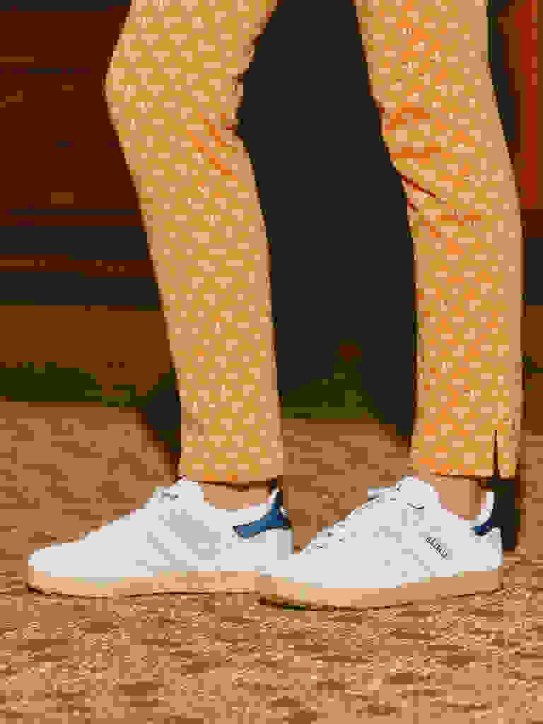 Na obrázku je vidět spodní část těla v vzorovaných oranžových kalhotách a bílých teniskách na dřevěné podlaze.