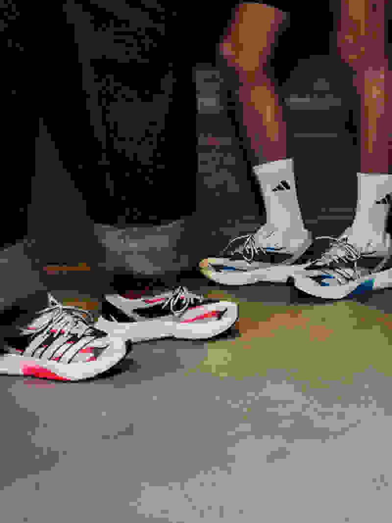 The image shows a pair of athletic shoes, likely sneakers, on a concrete or tiled floor. The shoes appear to be a mix of white, blue, and pink colors, and the person wearing them is wearing black pants and white socks.