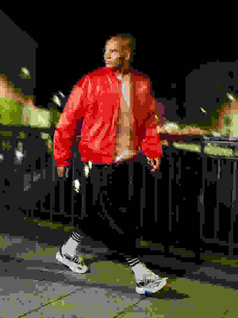 A person wearing a bright red jacket stands on a dimly lit street, with blurred buildings and structures visible in the background.