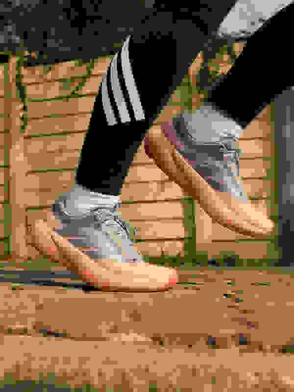 The image shows a pair of gray and orange athletic shoes with three white stripes on the side, standing on a paved surface in front of a wooden fence.