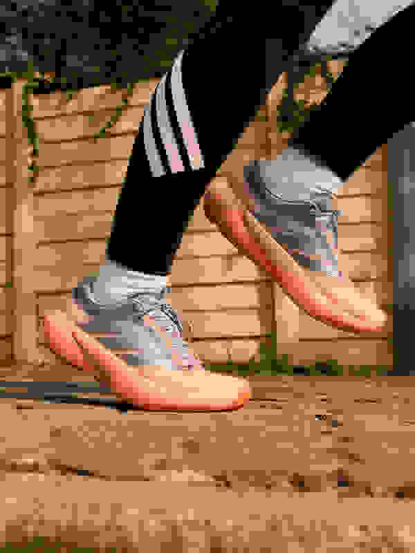 The image shows a pair of grey and orange athletic shoes on a person's feet, standing on a paved surface with a wooden fence in the background.