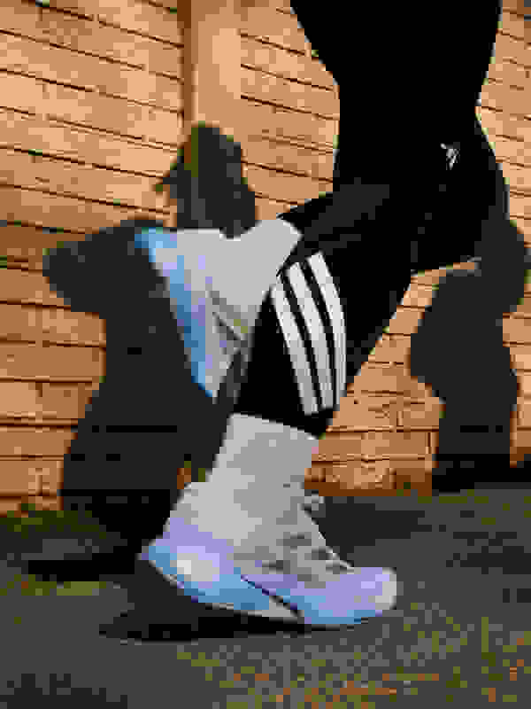 The image shows a person's legs and feet wearing a pair of white and blue athletic shoes against a brick wall background.