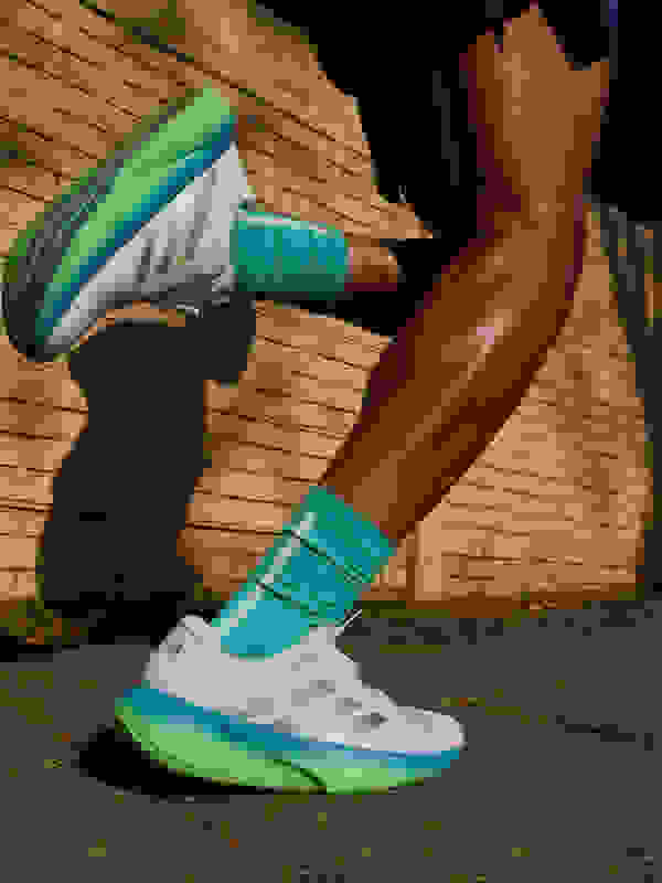 The image shows a person's lower body wearing a pair of green and white athletic shoes, standing on a brick wall with a shadowy background.