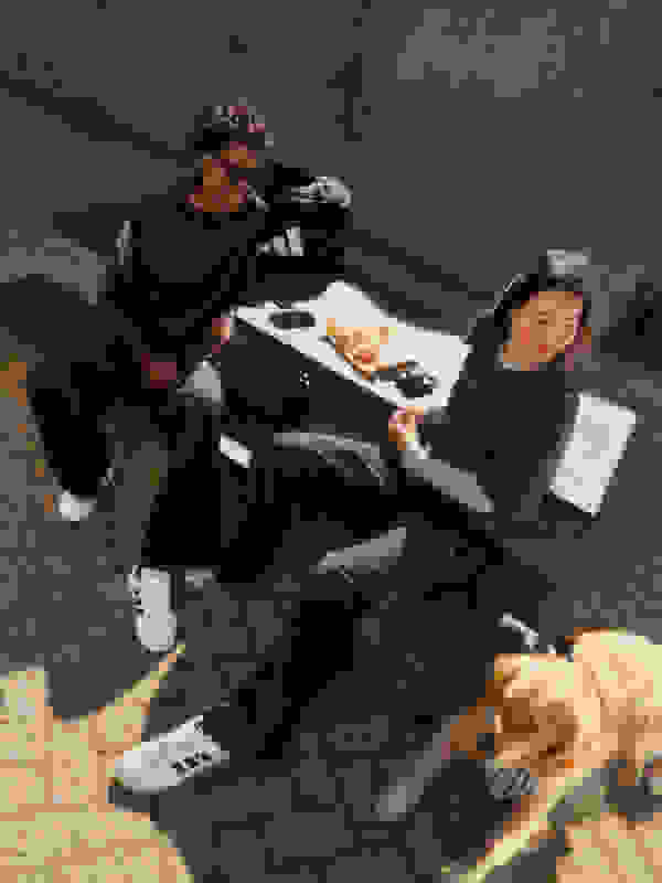 Twee individuen, een man en een vrouw, zitten op de grond in een stedelijke omgeving, met een hond in de buurt. De achtergrond lijkt een betonnen muur of structuur te zijn.