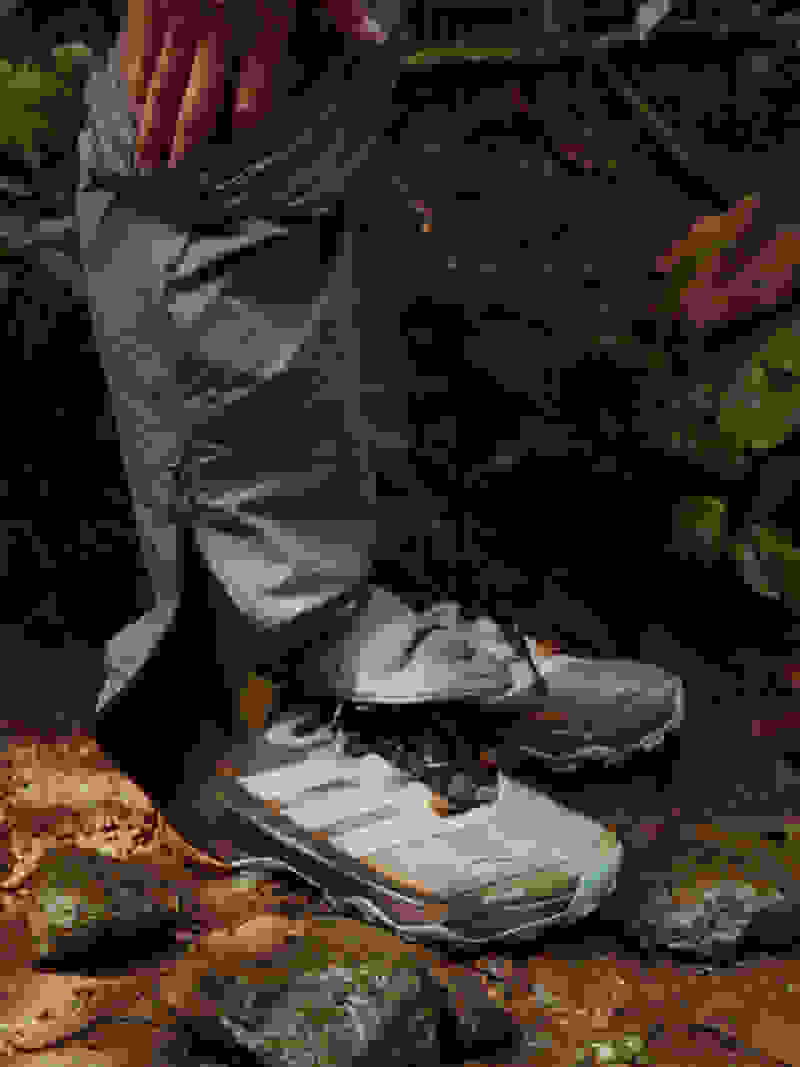 The image shows a person's lower body wearing hiking boots and trousers, standing on a rocky, forested path.